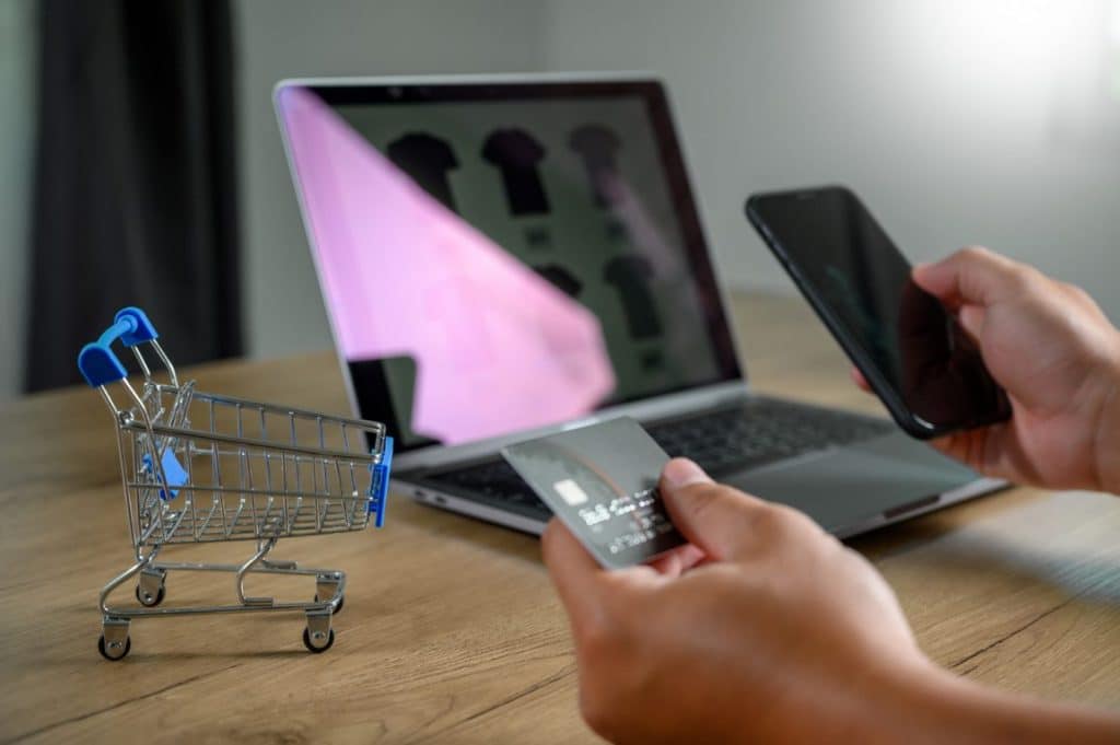 Persona con tarjeta y móvil en sus mano mirando un portátil y con un pequeño carro de compra a la derecha